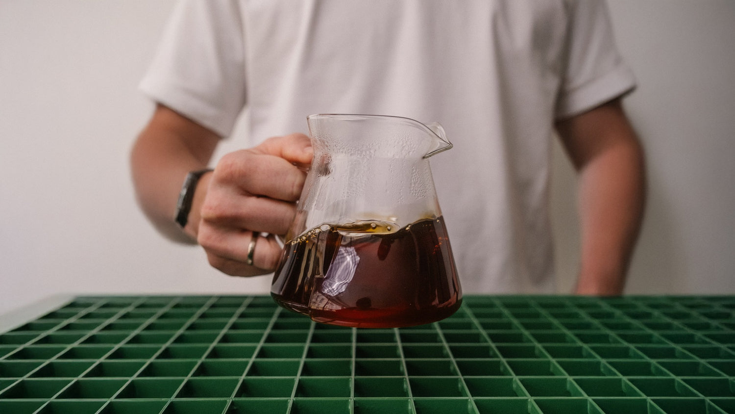 Hand hält gefüllte Kaffeekanne aus Glas ins Bild, darunter grünes Gitter